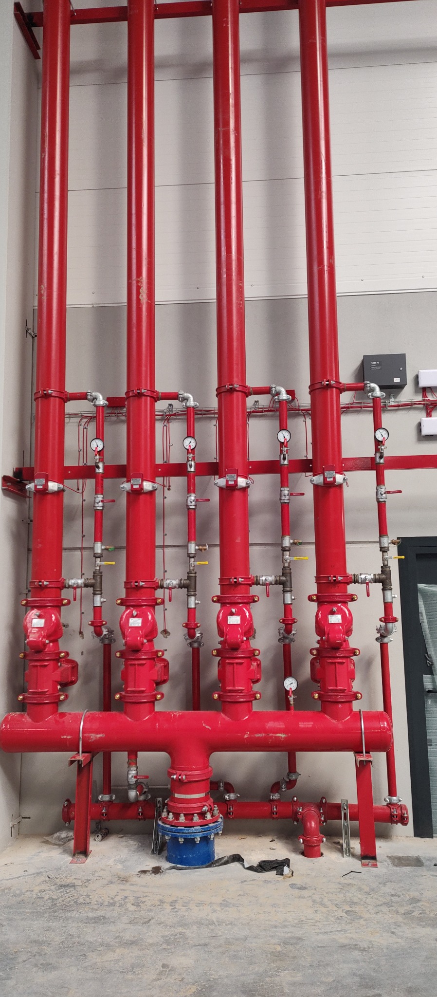 Czerwone rury pionowe i poziome instalacji przeciwpożarowej z zaworami, manometrami i niebieskim elementem łączeniowym u dołu, zamontowane przy szarej ścianie w hali.