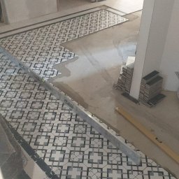 Bau Consult Vitalii Sliusarenko - Układanie wzorzystej terakoty na podłodze z użyciem poziomicy, widoczne zapasy płytek i świeży klej. Częściowo ułożona podłoga w trakcie prac wykończeniowych.