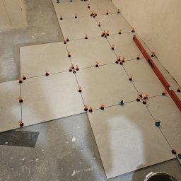 Bau Consult Vitalii Sliusarenko - Układanie jasnych płytek podłogowych z użyciem poziomicy i systemu poziomowania; widoczny fragment ściany i wiadro z zaprawą.