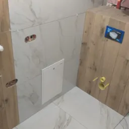 Wykończona łazienka z płytkami imitującymi drewno i marmur, widoczne otwory na gniazdka i rewizja. Użyto białych krzyżyków dystansowych.