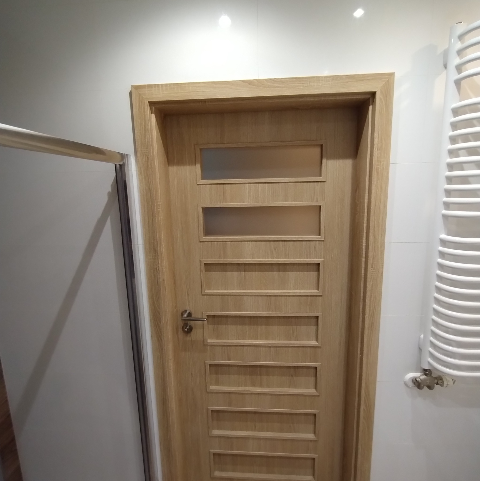 Jasna, drewnopodobna ościeżnica z przeszklonymi elementami w łazience. Widoczny fragment kabiny prysznicowej i grzejnik drabinkowy. Nowoczesny design.