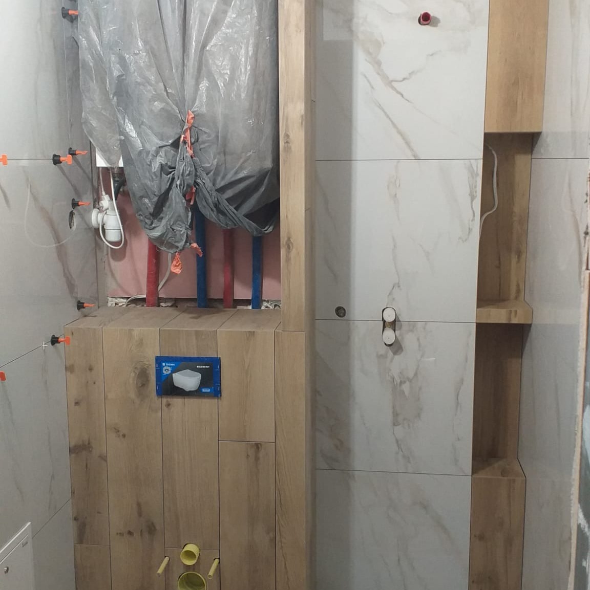 Montaż płytek w łazience z widoczną instalacją podtynkową. Częściowo zakryta folią. Użyto płytek imitujących drewno i marmur.