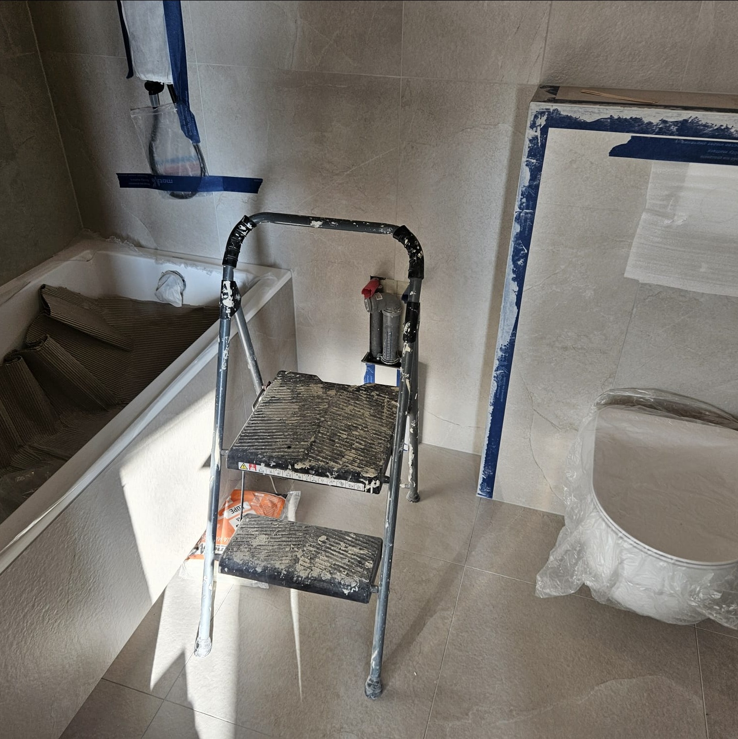 Remont łazienki: drabina malarska, zabezpieczona wanna, płyty do obłożenia ścian, wiadro z farbą w folii. Etap przygotowań do malowania ścian.