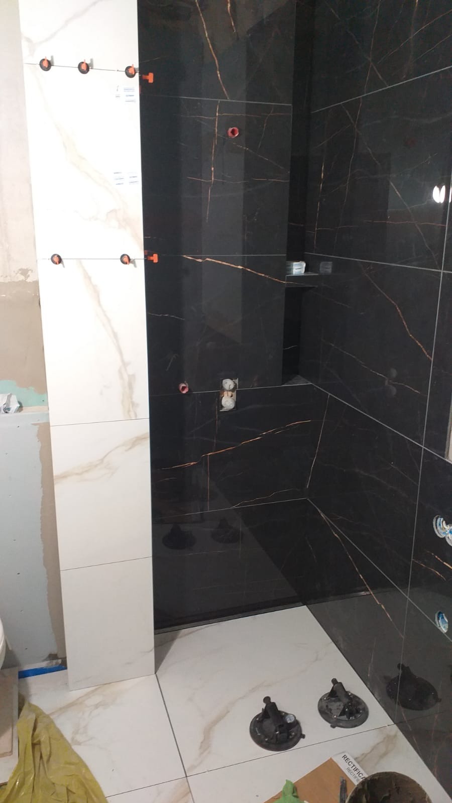 Układanie płytek w łazience: czarne i białe płytki z żyłkowaniem, system poziomowania płytek, przyssawki do płytek na podłodze. Widoczne gniazdka elektryczne.