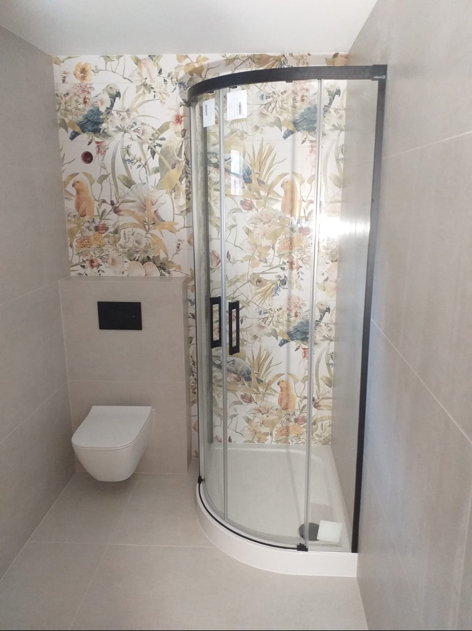 Nowoczesna łazienka z kabiną prysznicową z czarnymi ramami i toaletą. Ściany wyłożone płytkami z motywem kwiatowym i papug. Jasna, przestronna aranżacja.