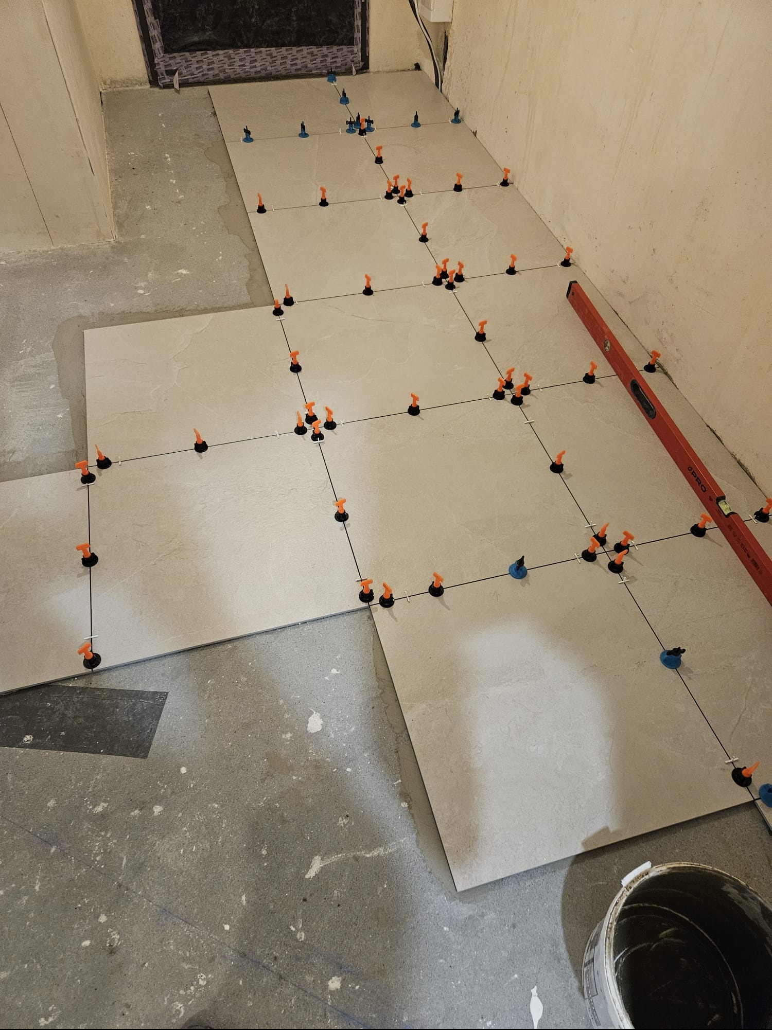 Układanie jasnych płytek podłogowych z użyciem poziomicy i systemu poziomowania; widoczny fragment ściany i wiadro z zaprawą.