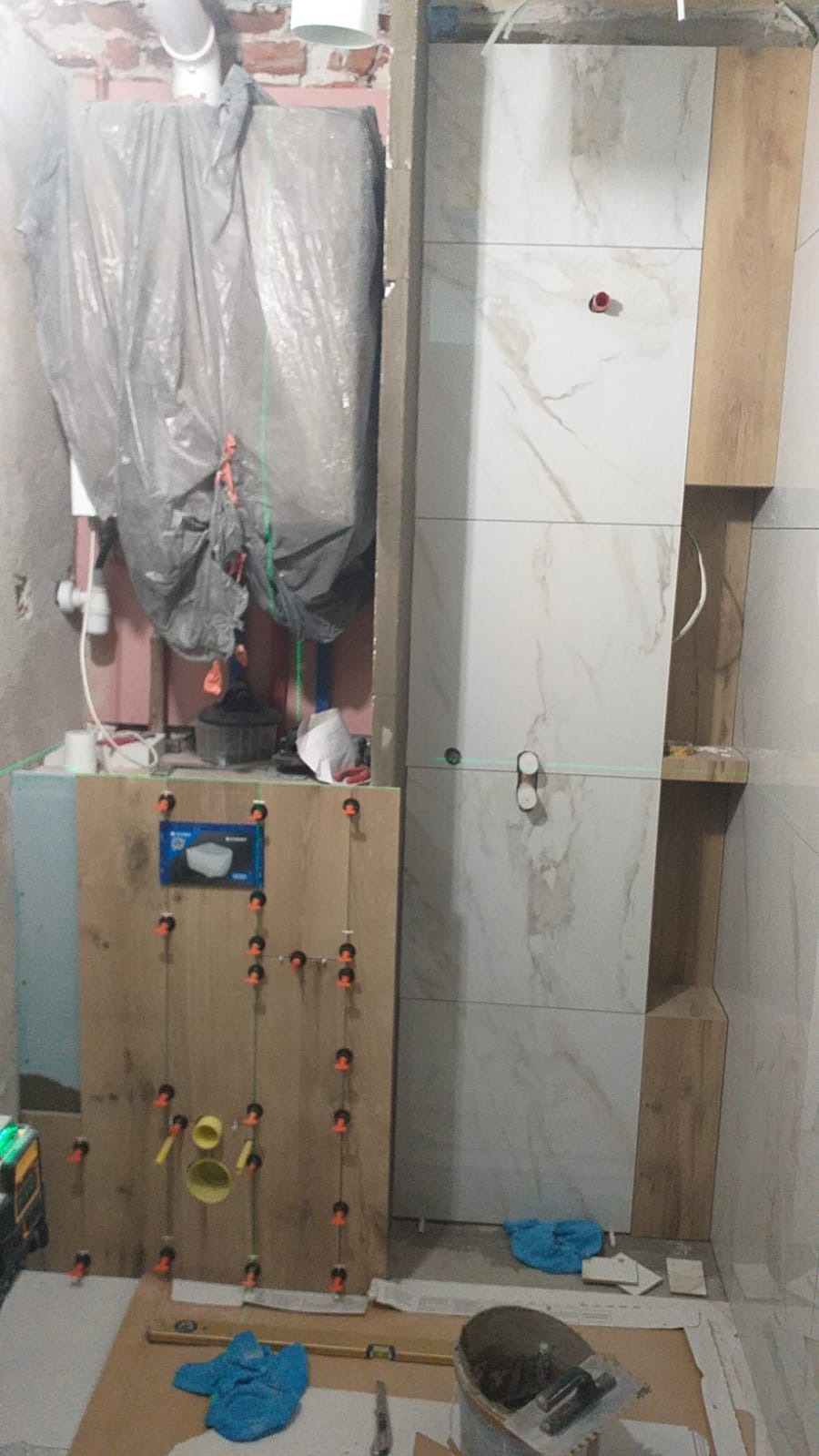 Remont łazienki: montaż płytek imitujących marmur na ścianie z ukrytą instalacją i stelażem WC. Widoczne narzędzia i poziomica laserowa.