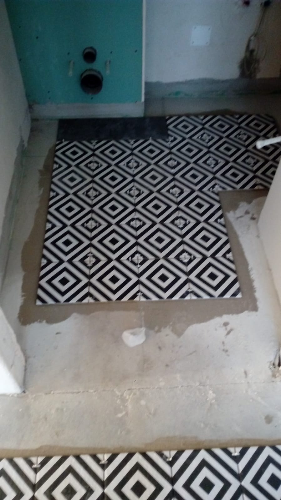 Układanie czarno-białych płytek podłogowych w łazience, widoczne rury odpływowe w ścianie, etap prac wykończeniowych. Geometryczny wzór płytek.