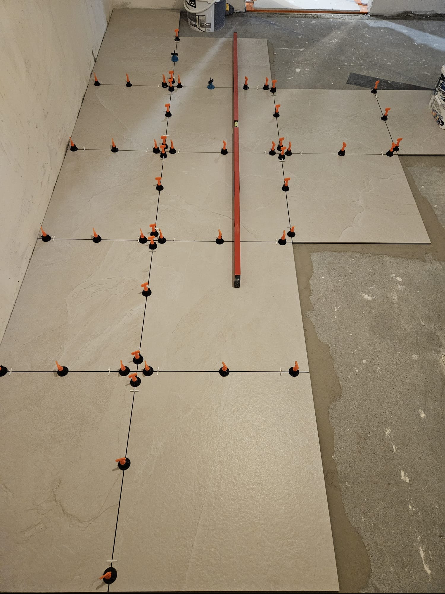 Układanie płytek podłogowych z użyciem systemu poziomowania; widoczny poziomica i wiadro z zaprawą. Jasne płytki z subtelnym wzorem, czarne klipsy i pomarańczowe kliny.