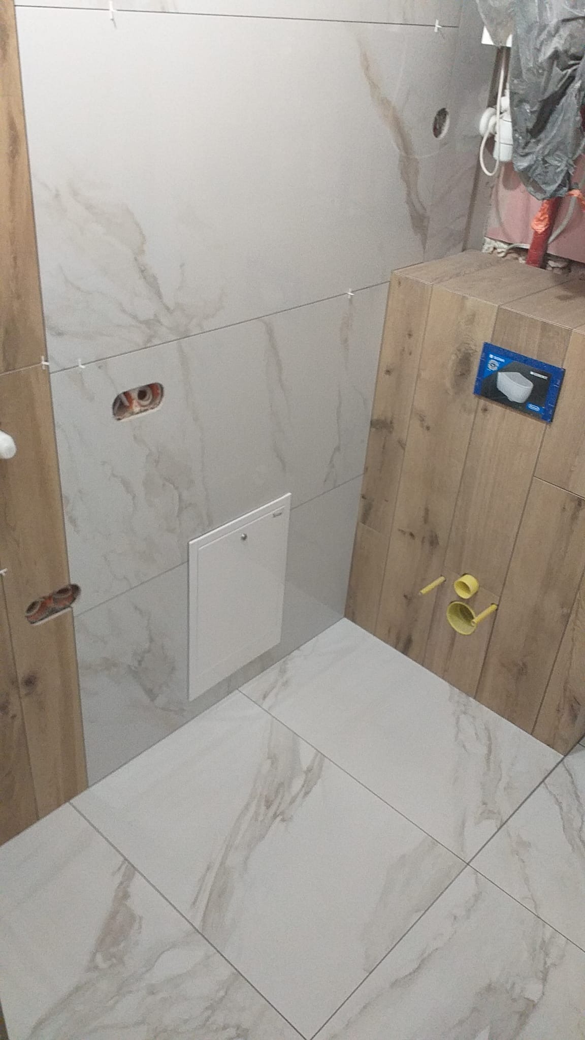 Wykończona łazienka z płytkami imitującymi drewno i marmur, widoczne otwory na gniazdka i rewizja. Użyto białych krzyżyków dystansowych.