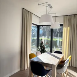 Jasne, kremowe zasłony w nowoczesnym wnętrzu jadalni z drewnianym stołem i kolorowymi krzesłami, widok na zewnątrz przez okno z choinką.