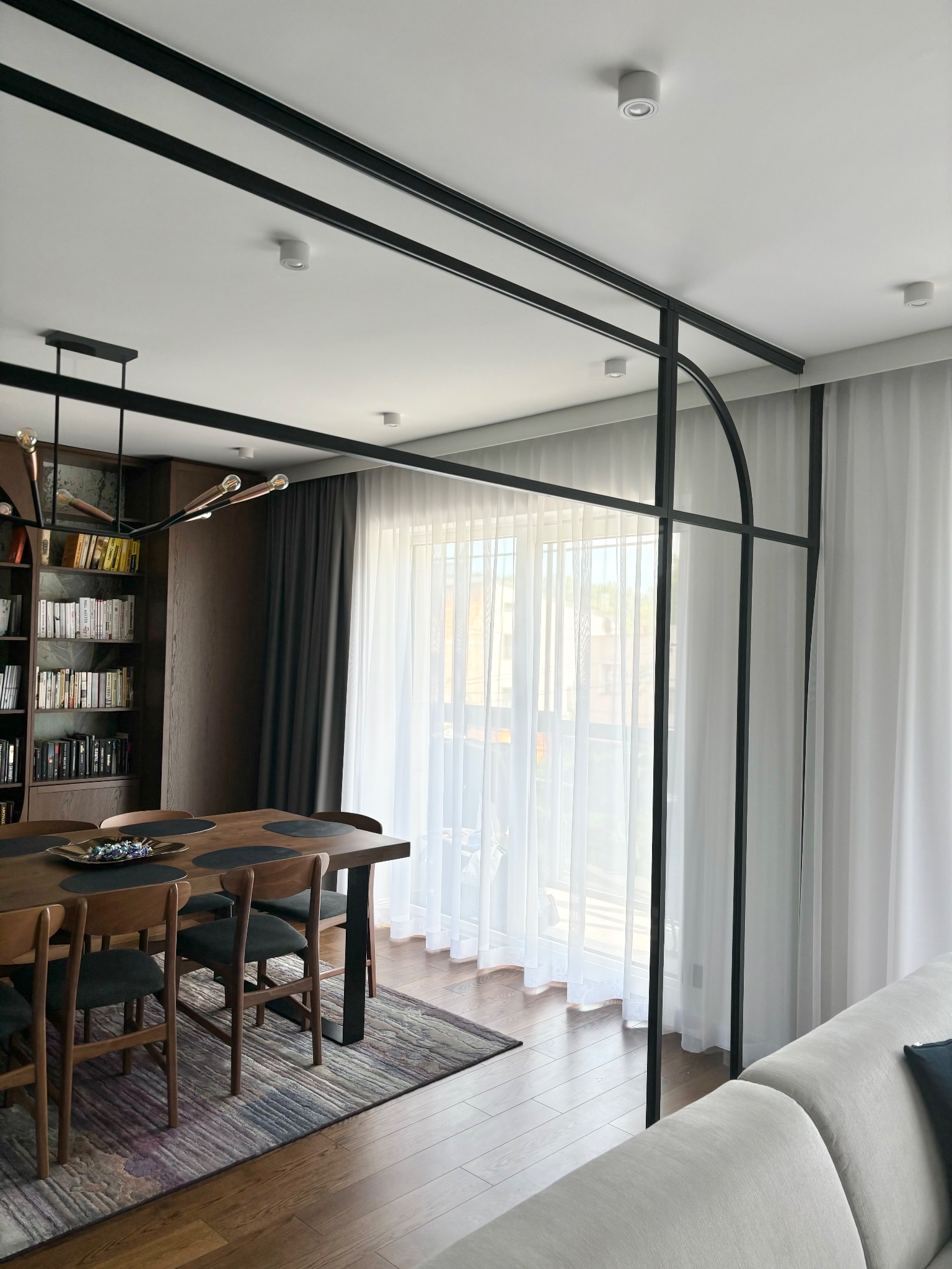 Jasne wnętrze jadalni z dużym oknem, przezroczystymi białymi zasłonami i ciemnoszarymi zasłonami po lewej stronie okna, dzielonym czarną metalową konstrukcją na wzór okna, drewnianym stołem...
