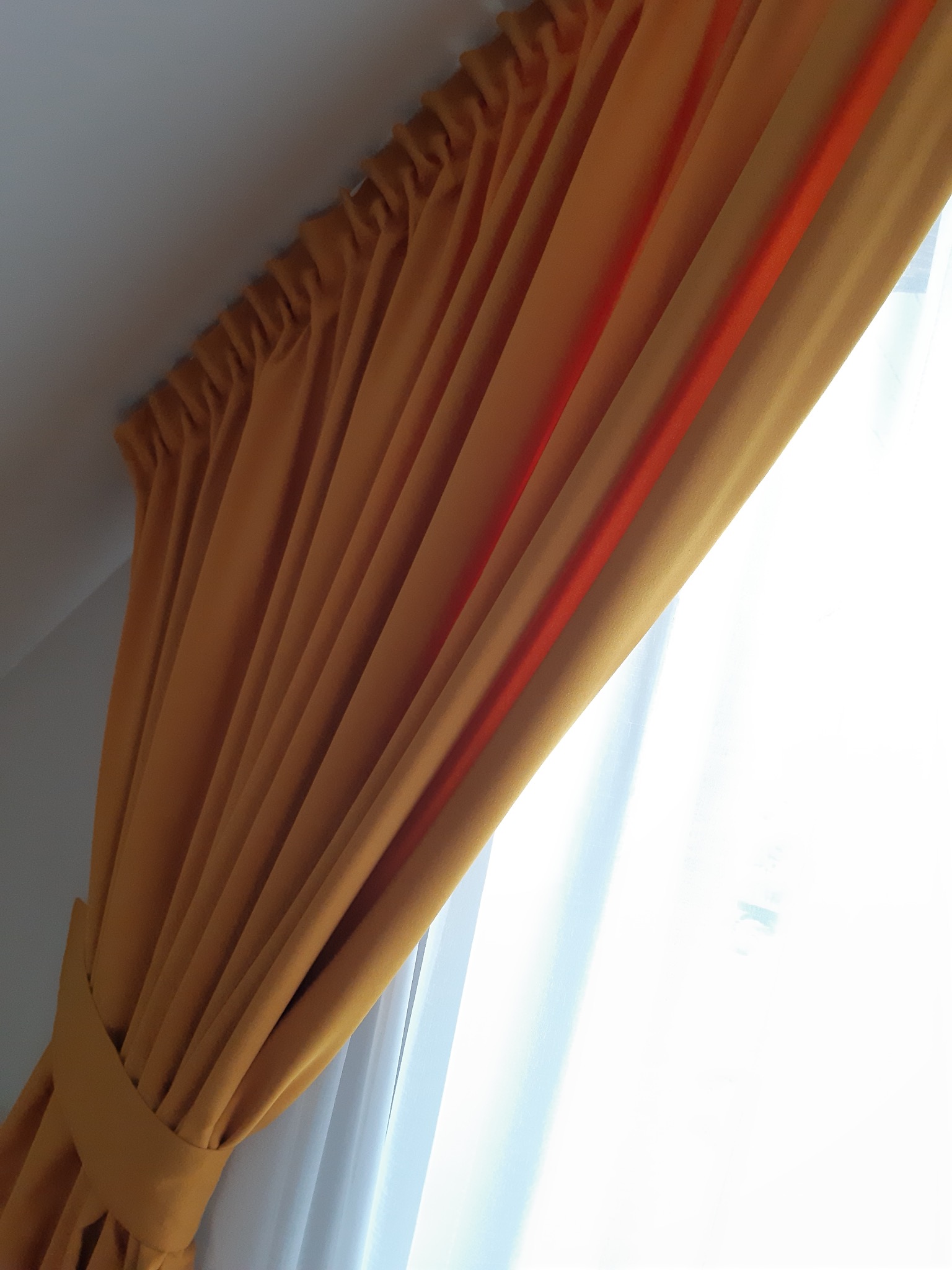 Złota zasłona z pomarańczowym akcentem, upięta pasem materiału, z widocznym fragmentem białej firanki i jasnym światłem wpadającym przez okno.