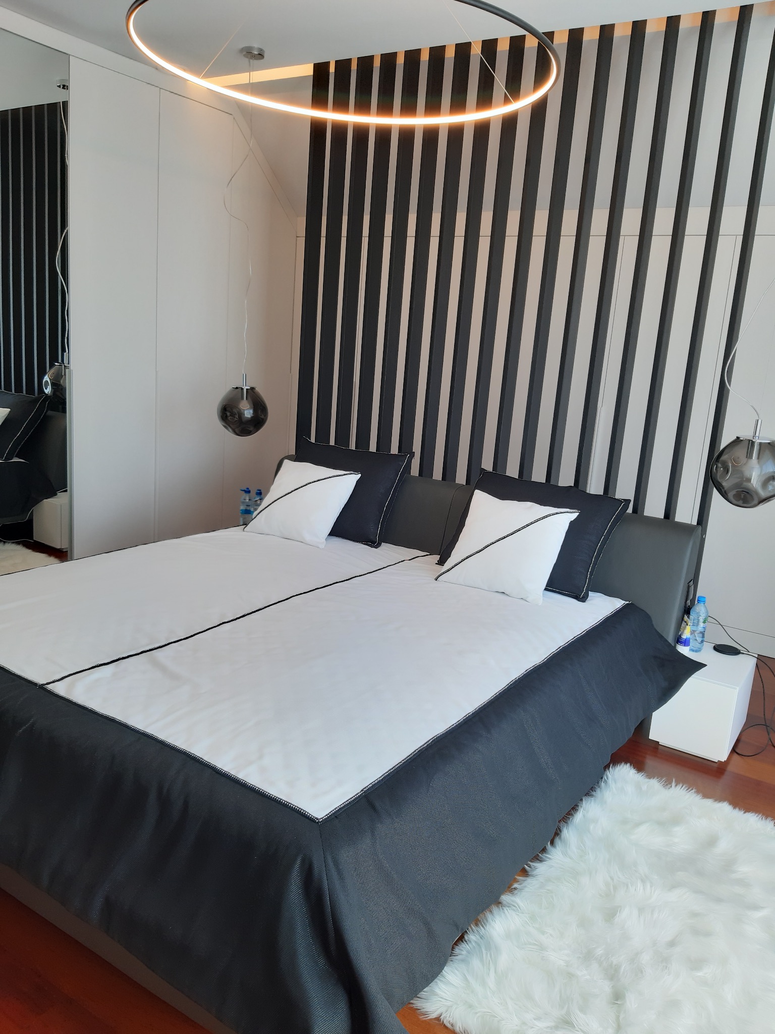 Nowoczesna sypialnia z łóżkiem dwuosobowym w bieli i czerni, dekoracyjna ściana z pionowych listew, okrągła lampa sufitowa i puszysty dywanik.