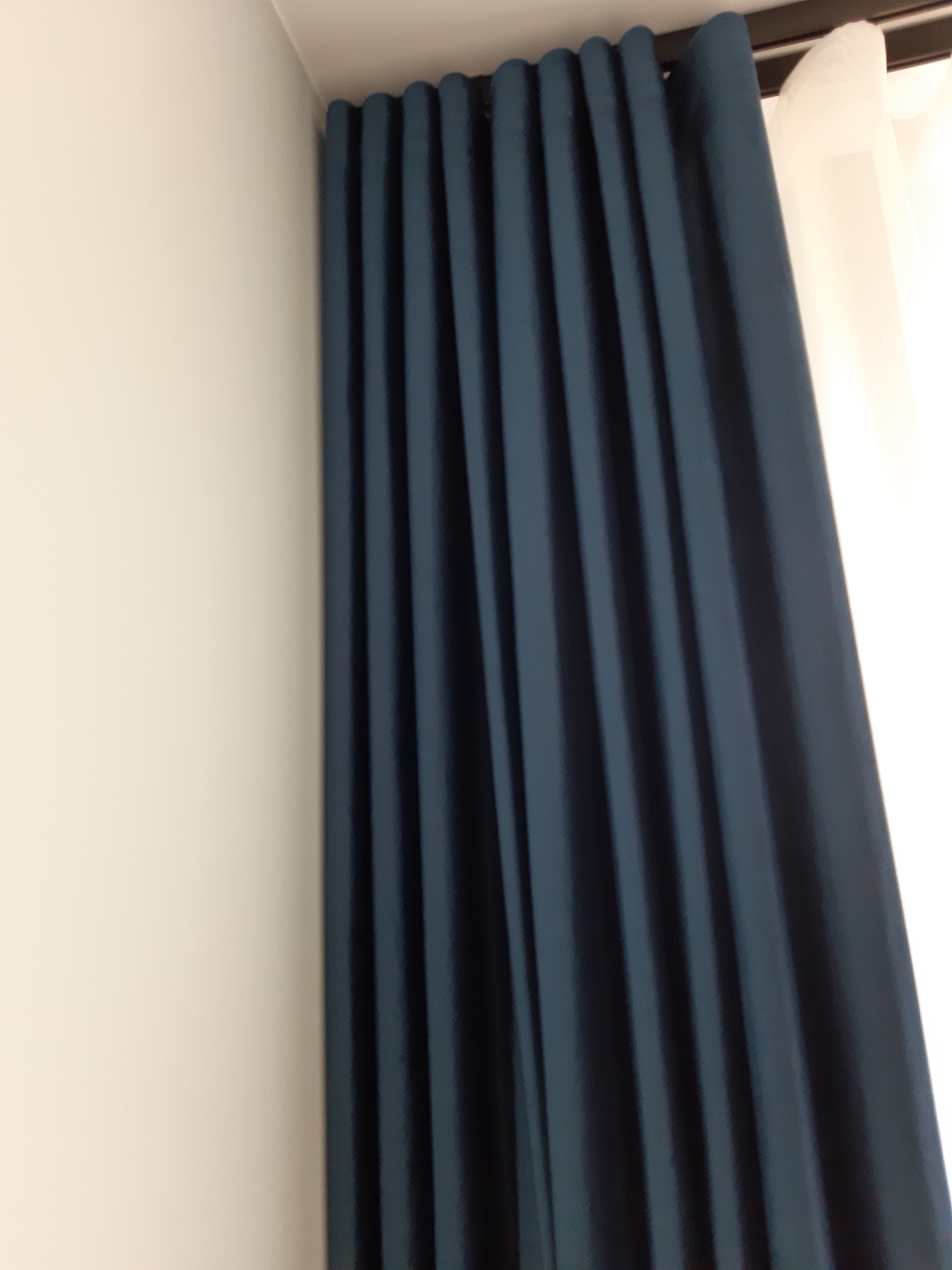 Długie, granatowe zasłony z regularnymi fałdami, zamontowane na czarnym karniszu, z fragmentem białej firanki po prawej stronie, przylegające do białej ściany.