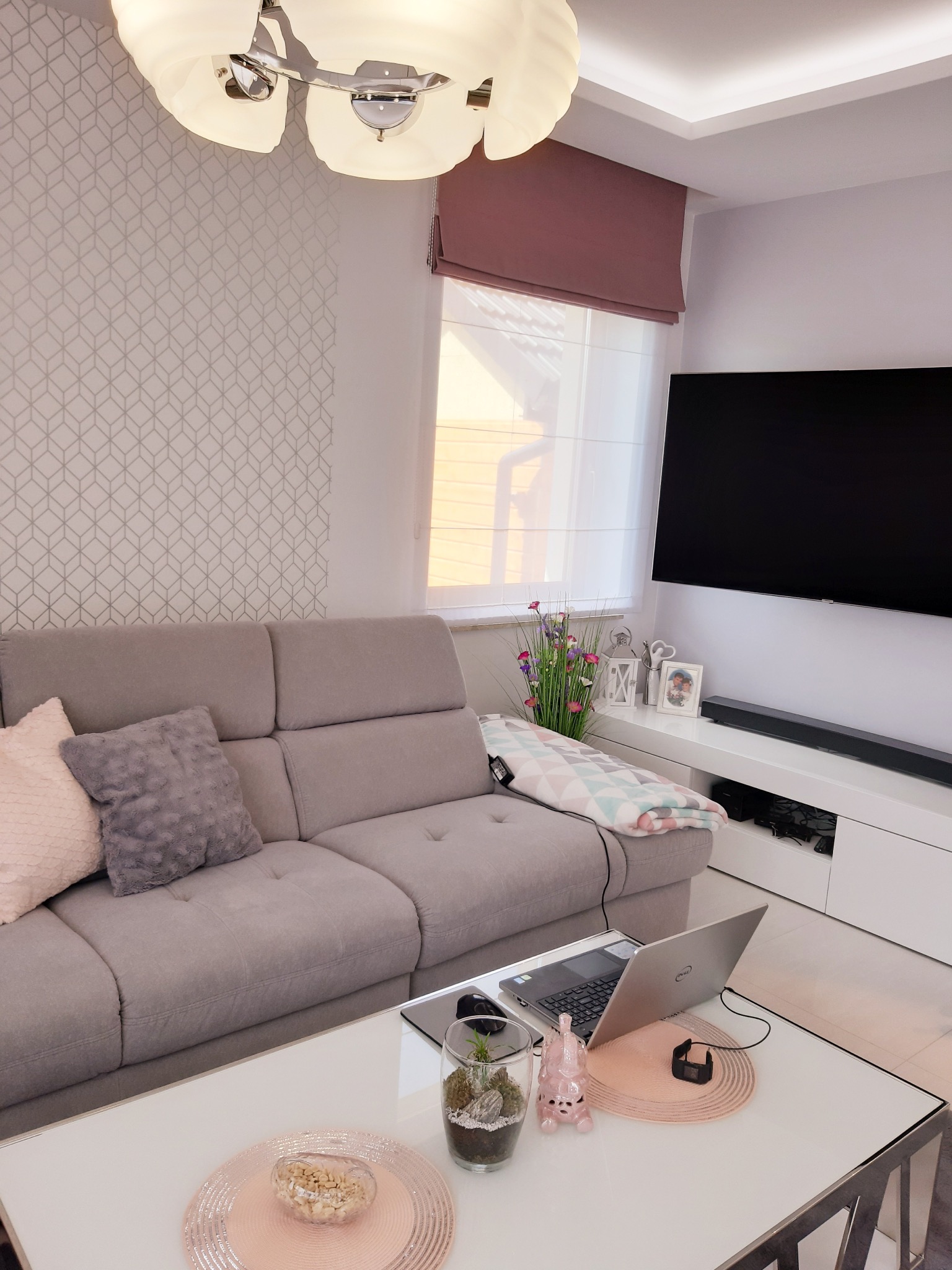 Nowoczesny salon z szarą sofą, laptopem na stoliku kawowym i różową roletą rzymską w oknie, uzupełniony geometryczną tapetą i dekoracyjnym oświetleniem.