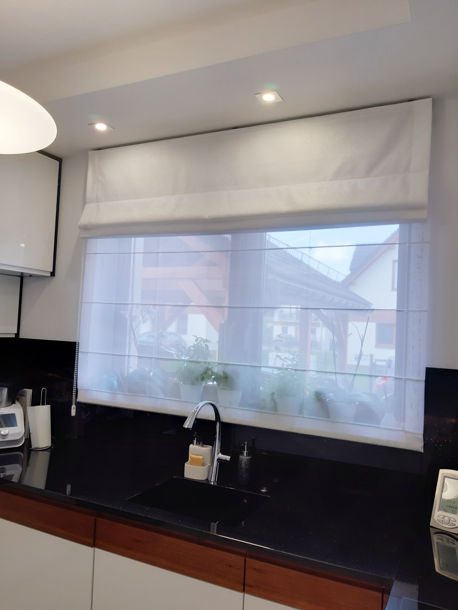 Biała roleta rzymska na oknie w kuchni z czarnym blatem, widok na ogród z roślinami doniczkowymi na parapecie.