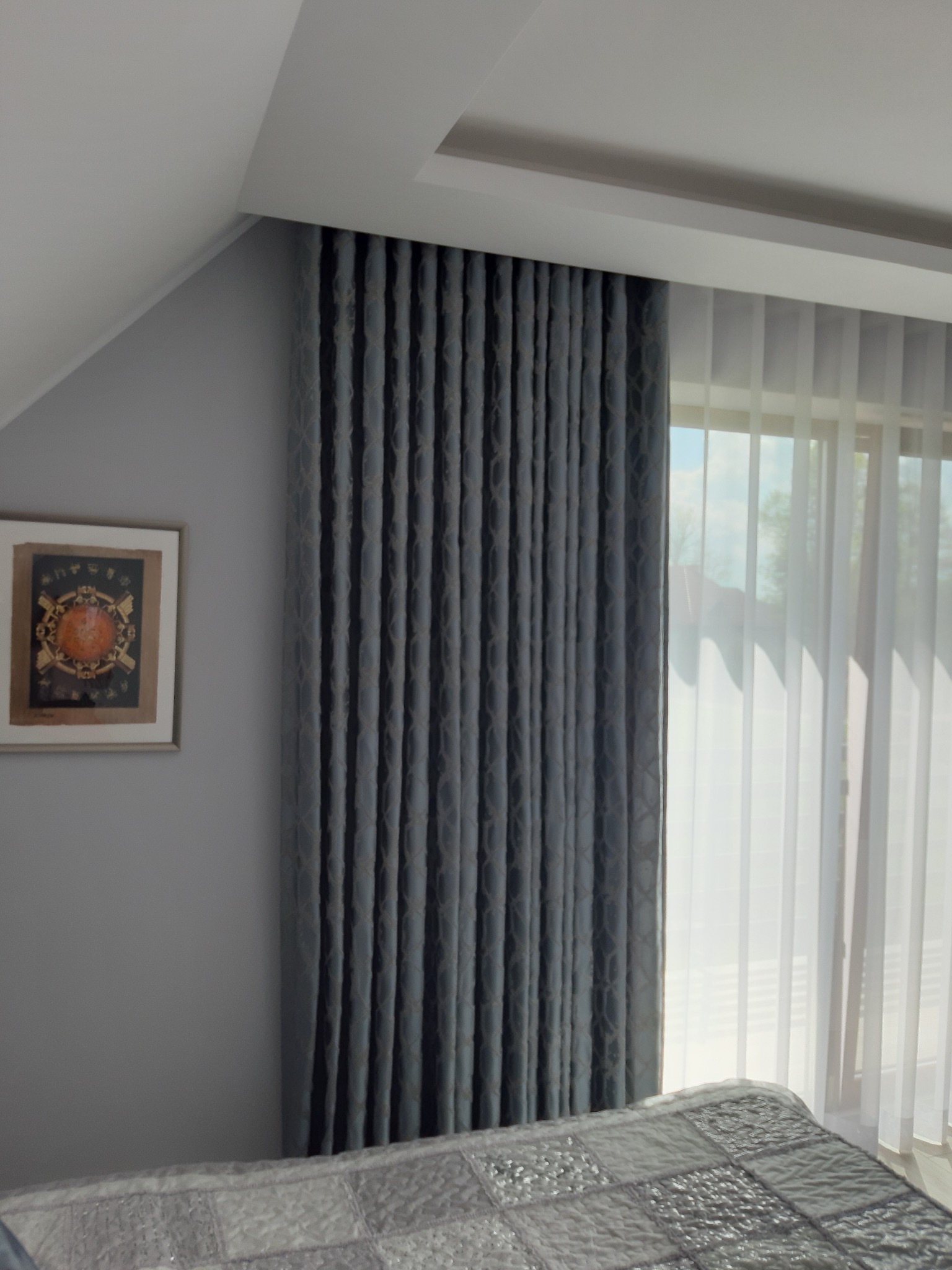 Zasłony w odcieniach szarości z delikatnym wzorem geometrycznym, upięte w regularne fałdy, wiszące obok okna z białymi, przejrzystymi firanami, fragment sypialni z łóżkiem i obrazem na ścianie.