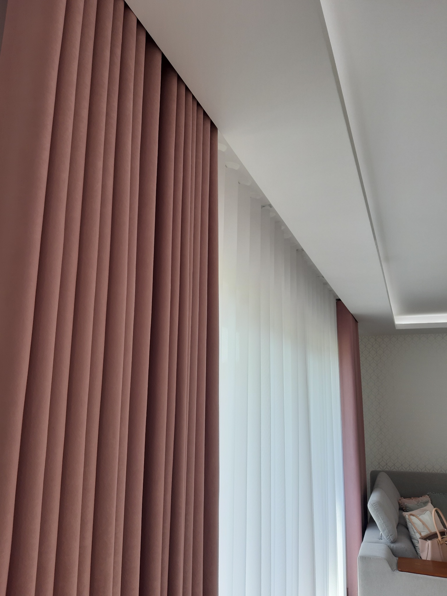 Ciemnoróżowe, grube zasłony z plisami, zestawione z białymi, pionowymi żaluzjami w nowoczesnym wnętrzu z szarą sofą w tle.