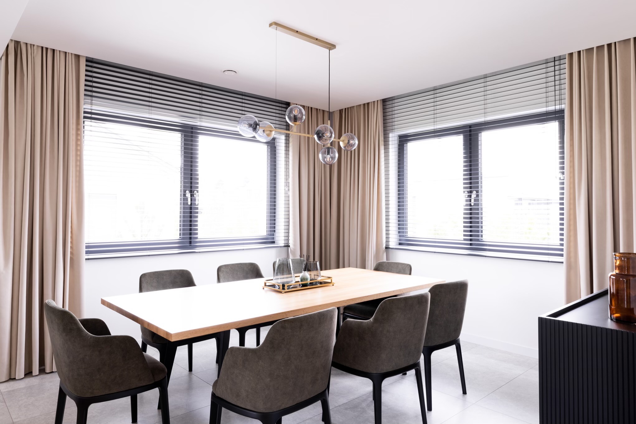 Jasne wnętrze jadalni z beżowymi zasłonami po bokach okien z żaluzjami, drewnianym stołem otoczonym szarymi krzesłami i nowoczesną lampą wiszącą z szklanymi kulami.
