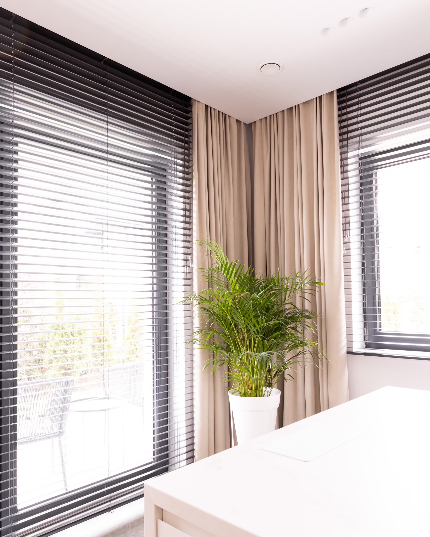 Jasne wnętrze z beżowymi zasłonami, czarnymi żaluzjami i zieloną rośliną doniczkową w białej osłonce, widok na balkon.