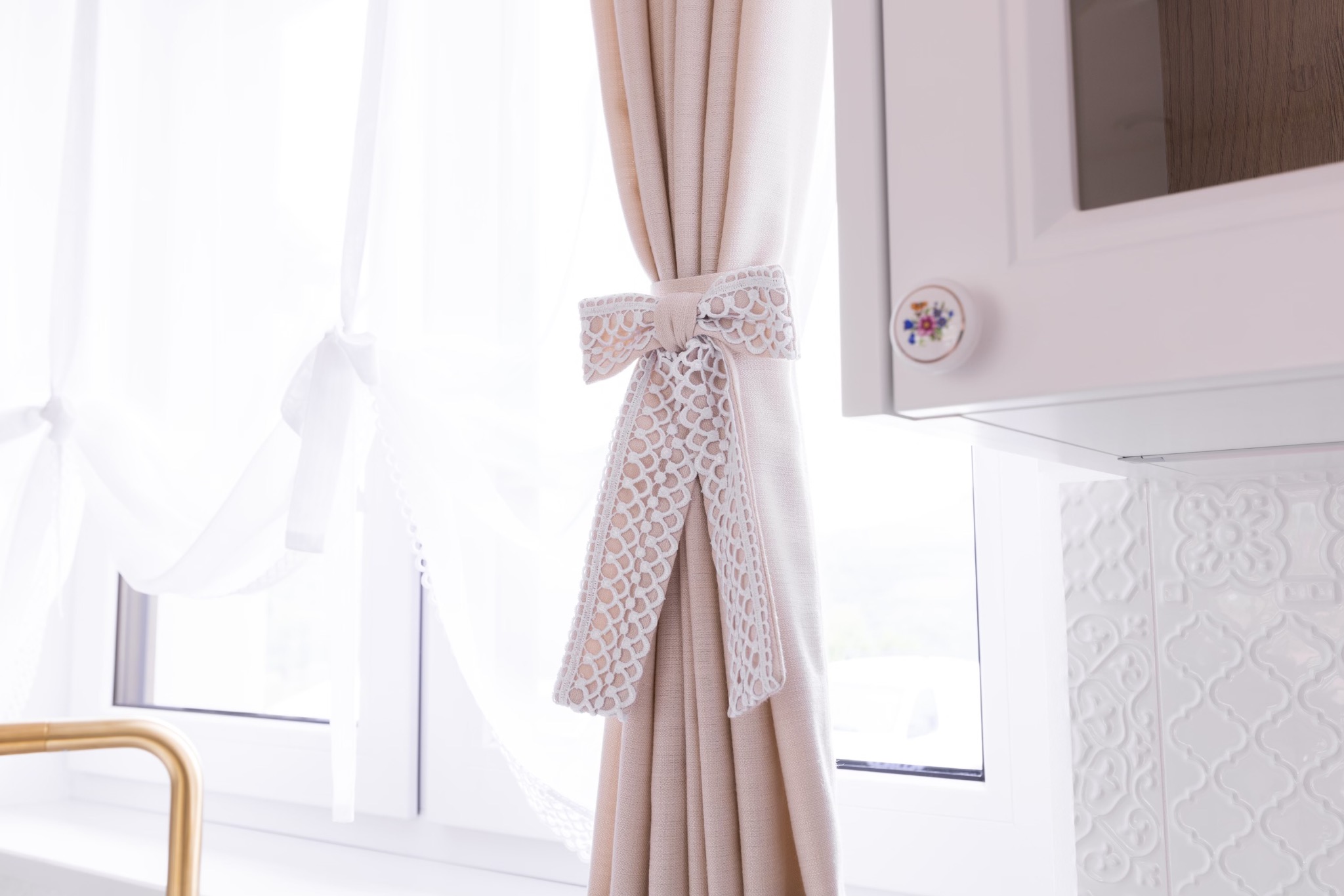 Beżowa zasłona upięta ozdobną, kremową wstążką z koronkowym wzorem na tle okna i białych mebli kuchennych z dekoracyjnym uchwytem.