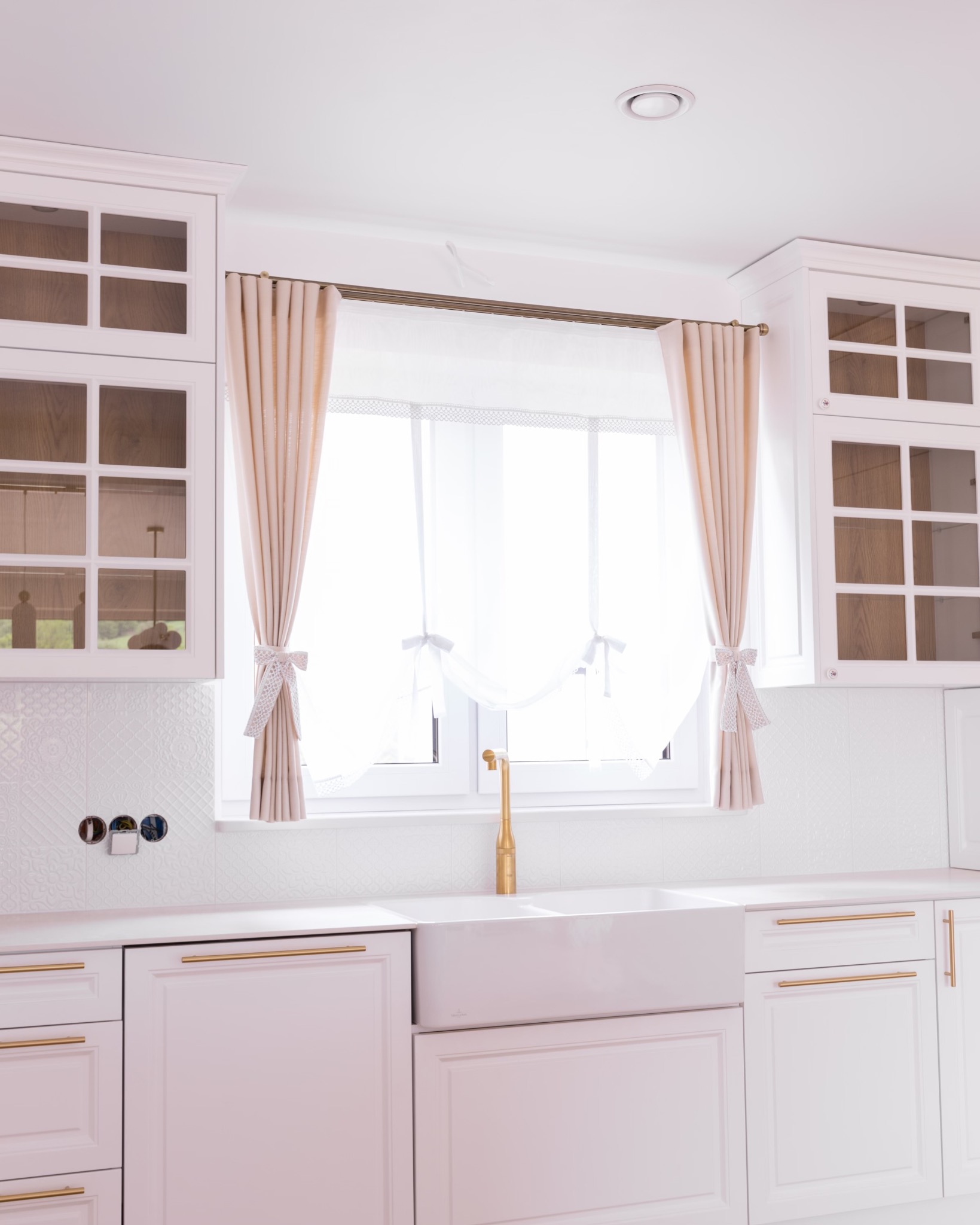 Jasna kuchnia w Krakowie z białymi szafkami, złotymi uchwytami i zlewem farmerskim, okno zdobią beżowe zasłony podwiązane ozdobnymi kokardami.