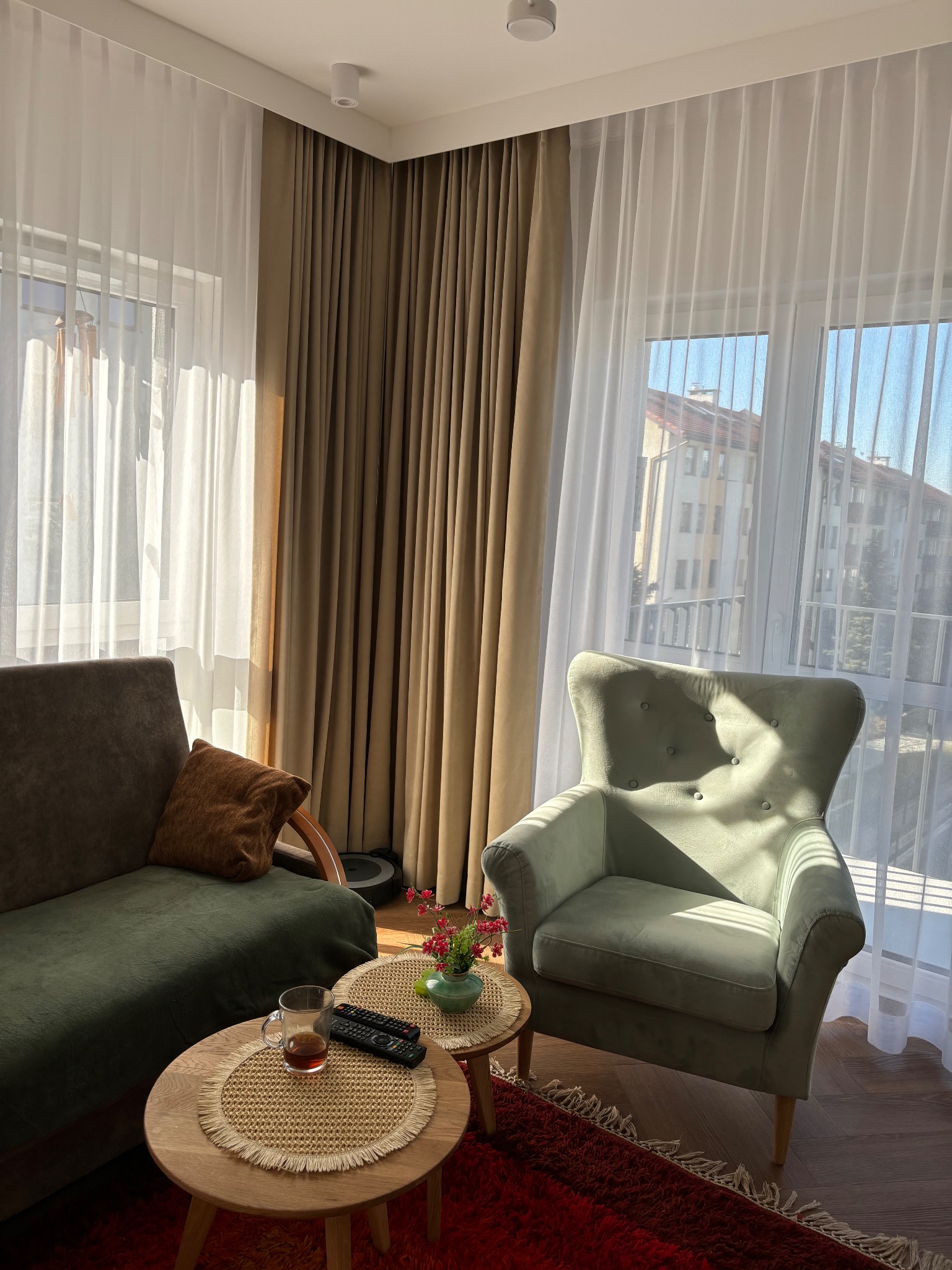 Jasne wnętrze pokoju dziennego z zielonym fotelem i sofą, okno przesłonięte białymi, prześwitującymi i brązowymi, zaciemniającymi zasłonami.