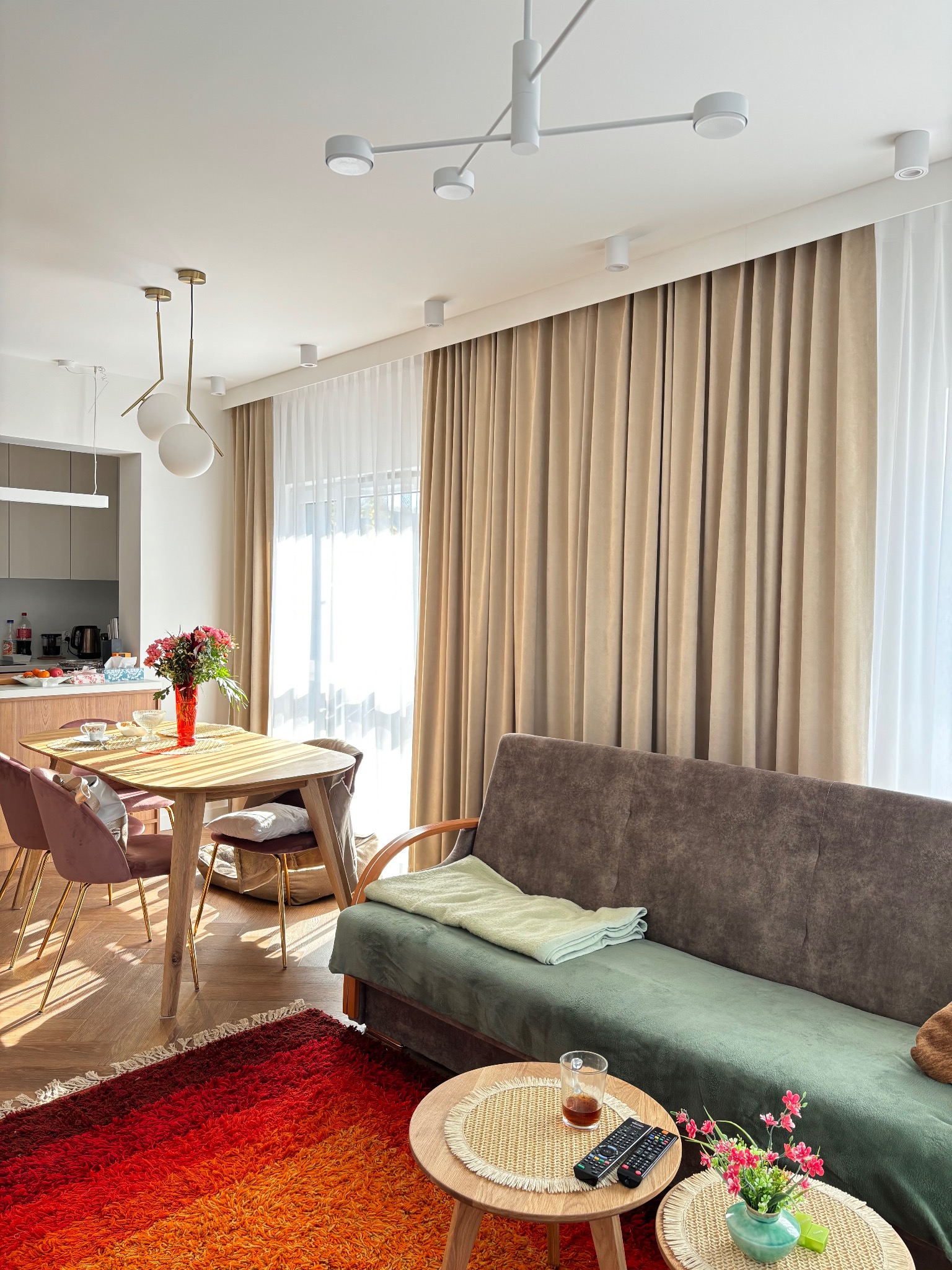 Beżowe i białe zasłony w jasnym salonie w Krakowie, widoczne z perspektywy dywanu w odcieniach czerwieni i pomarańczy, z drewnianym stołem i krzesłami w tle.