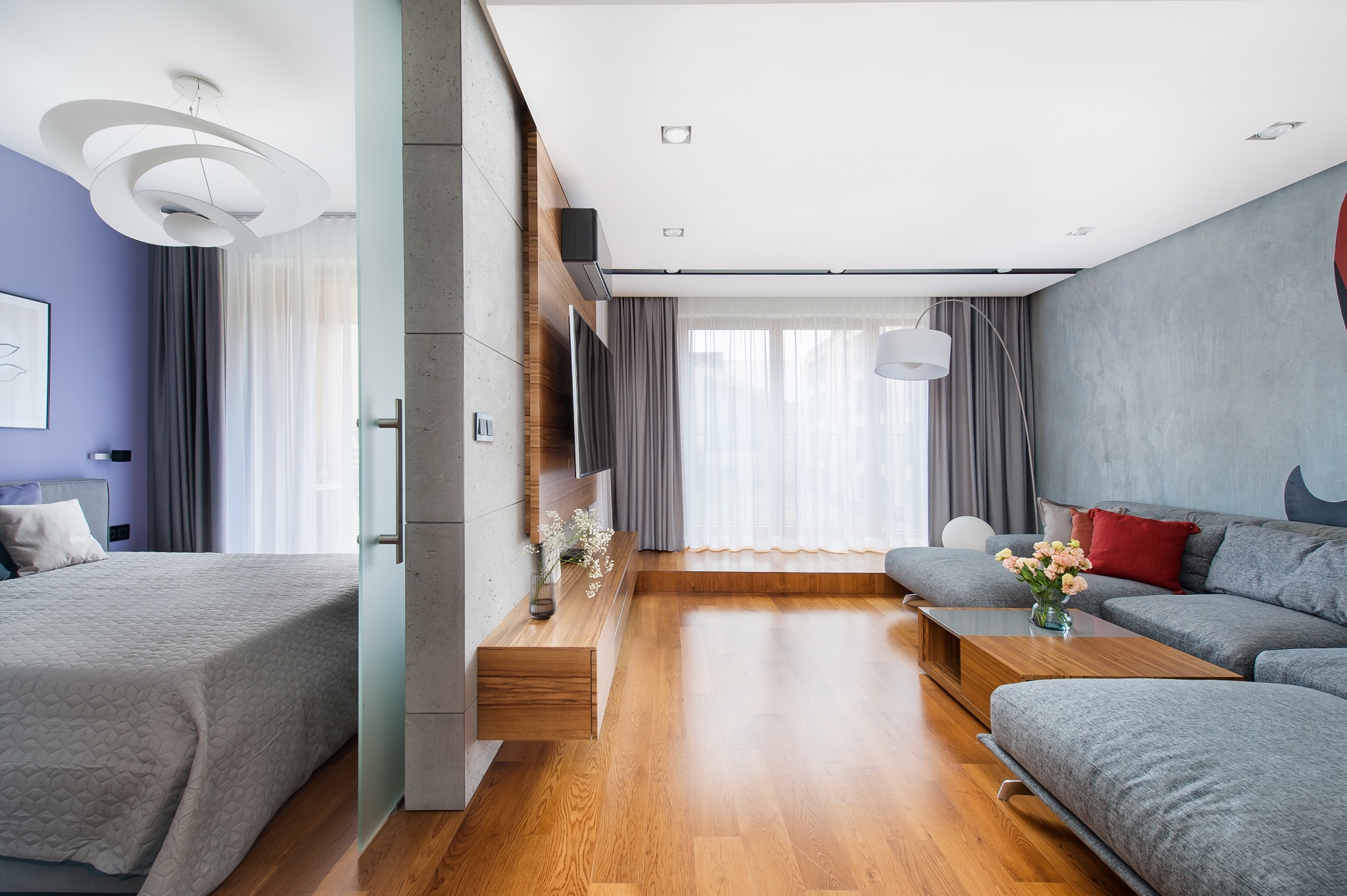 Nowoczesne wnętrze salonu z szarym narożnikiem, drewnianym parkietem i zasłonami w oknie balkonowym, widok na sypialnię przez szklane drzwi.