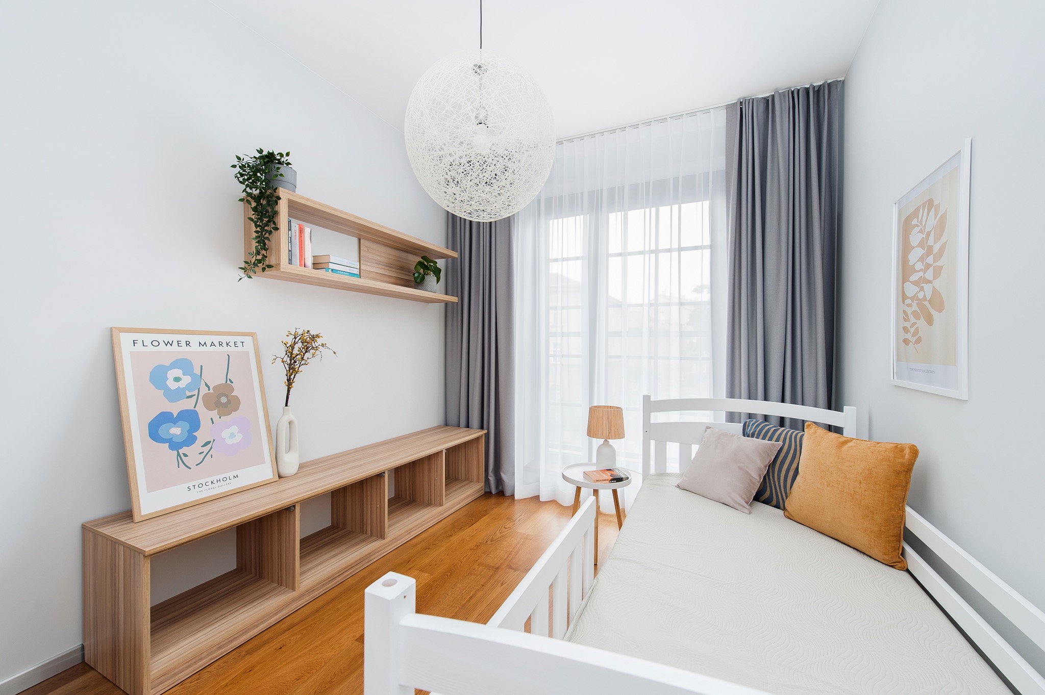 Jasny pokój z drewnianą podłogą, białym łóżkiem, szarą zasłoną i plakatem 'Flower Market' na ścianie, oświetlony kulistą lampą wiszącą.
