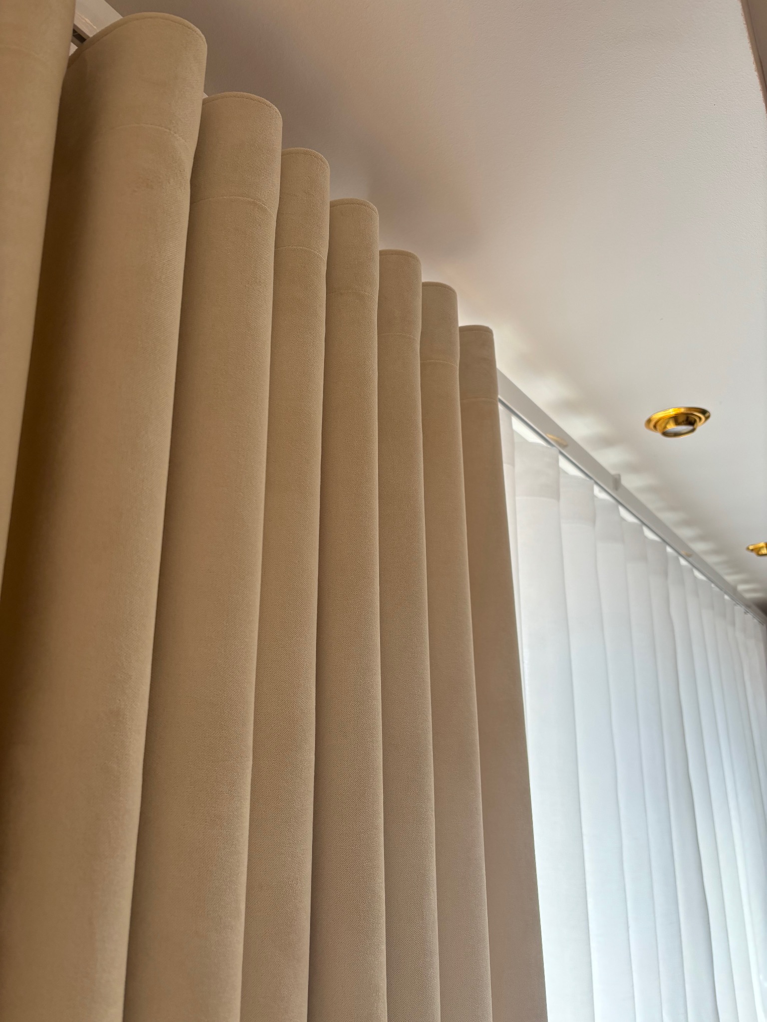 Beżowe, grube zasłony z widocznym systemem mocowania do sufitu, obok prześwitujące, białe, pionowe żaluzje.