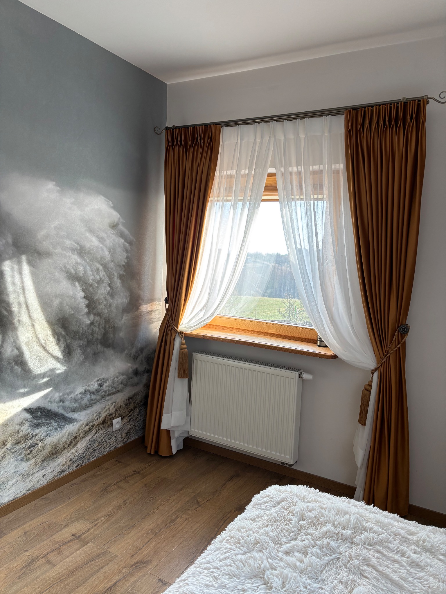 Okno z brązowo-białymi zasłonami związanymi ozdobnymi sznurami z frędzlami; na ścianie fototapeta z motywem wzburzonego morza.