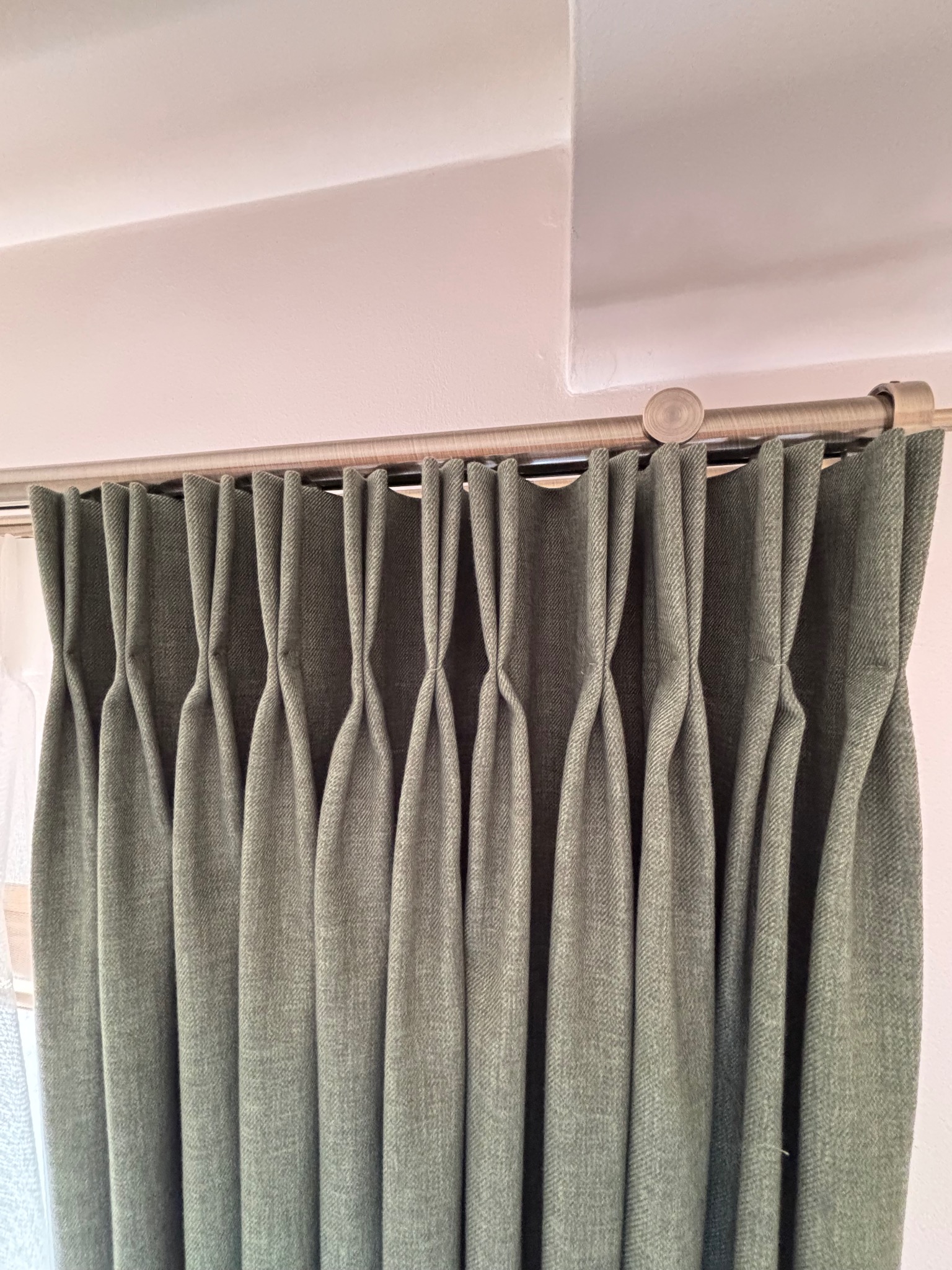 Zielone, grube zasłony z zakładkami, zawieszone na metalowym karniszu z drewnianymi zakończeniami, widoczne spod sufitu.