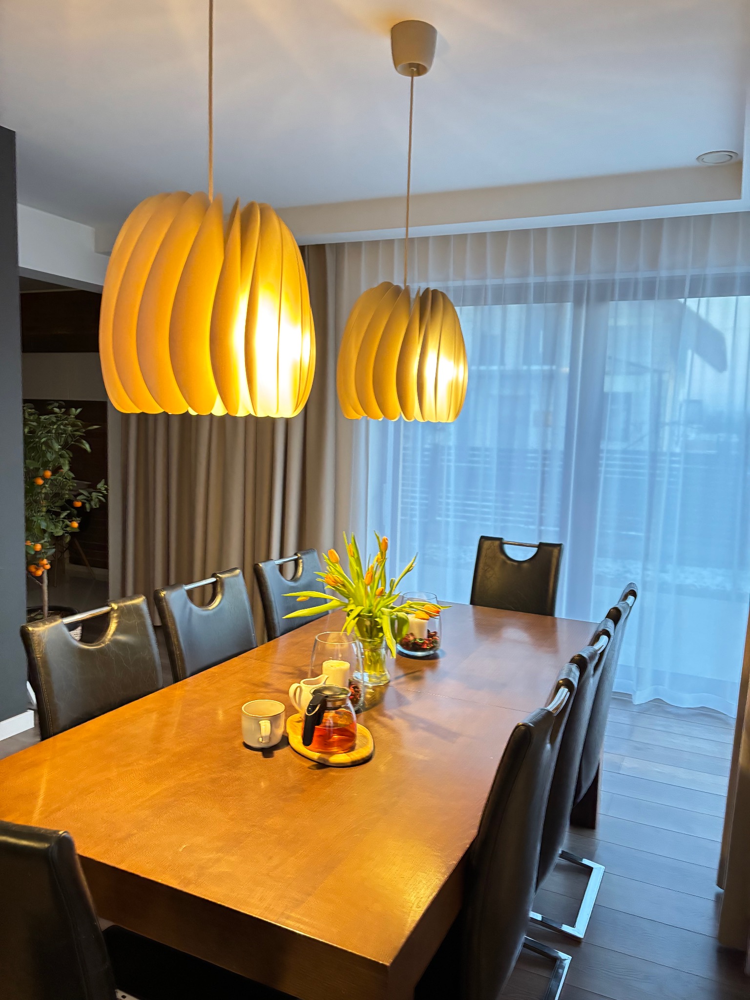 Jadalnia z długim drewnianym stołem, krzesłami obitymi skórą, tulipanami w wazonie i dwoma wiszącymi lampami o organicznym kształcie, oświetlona naturalnym światłem przez zasłony okienne.