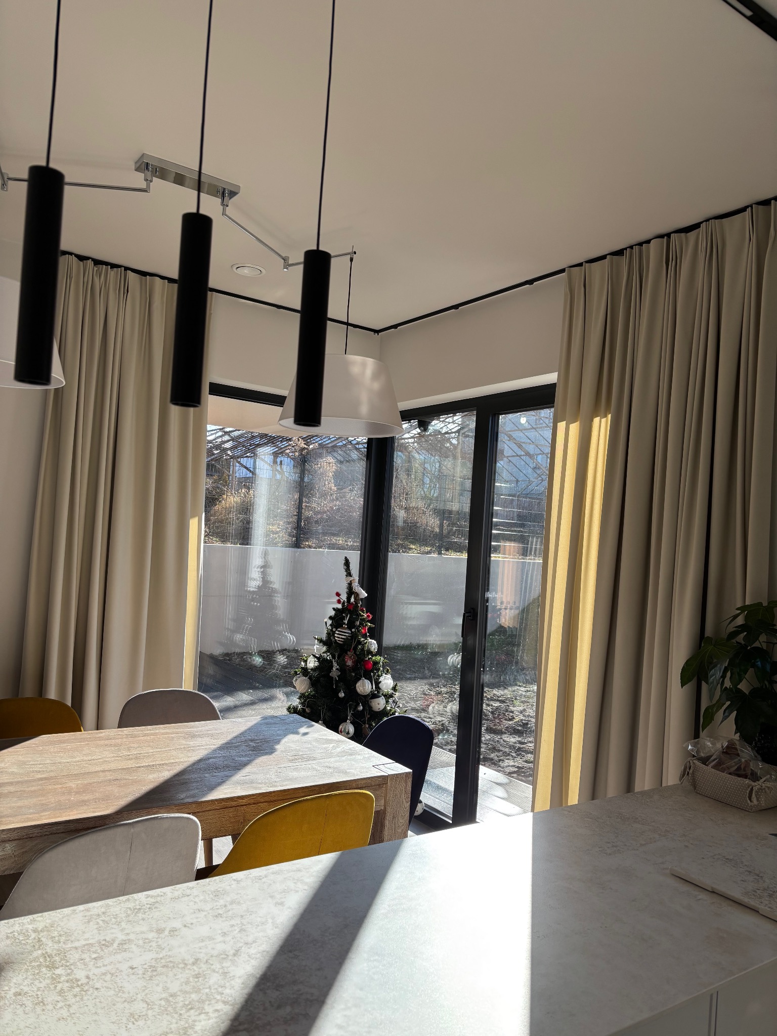 Jasne, beżowe zasłony w nowoczesnym wnętrzu z widokiem na ogród przez przeszklone drzwi, oświetlone przez wiszące lampy i padające promienie słońca, z choinką w tle.