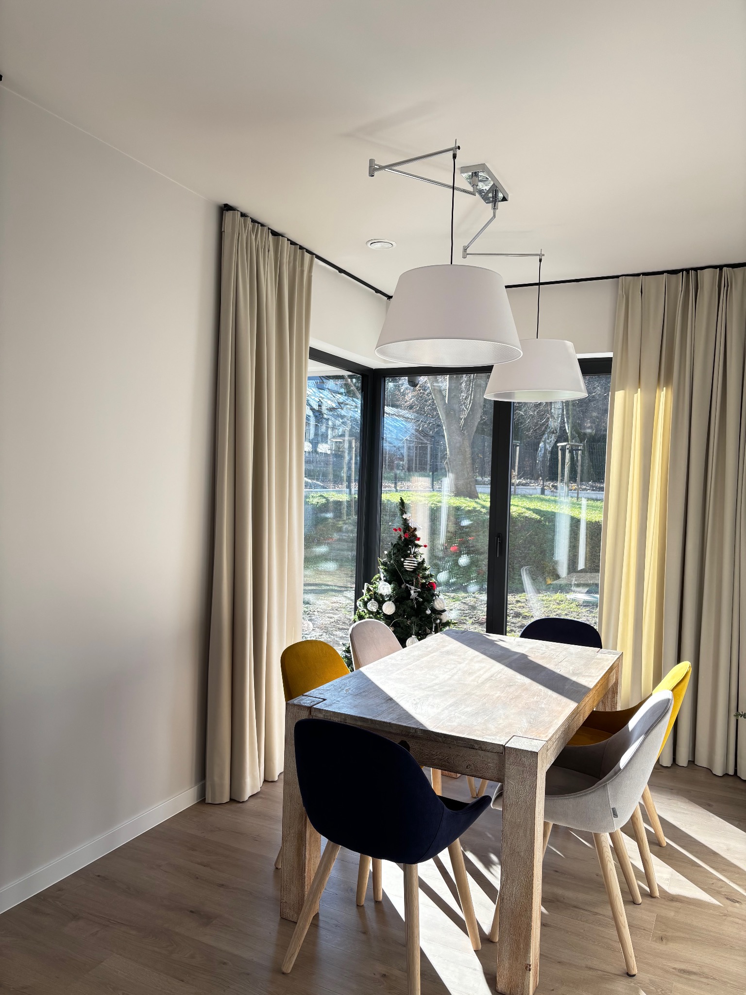Jasne, kremowe zasłony w nowoczesnym wnętrzu jadalni z drewnianym stołem i kolorowymi krzesłami, widok na zewnątrz przez okno z choinką.