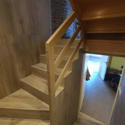 Firma ogolnobudowlana Jarzmik Kamil - Drewniane schody z balustradą w wykończonym wnętrzu, z widoczną okładziną ścienną imitującą drewno i fragmentem pomieszczenia za otwartymi drzwiami.