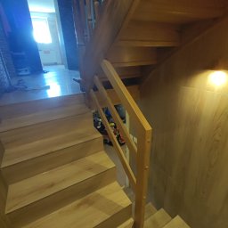 Firma ogolnobudowlana Jarzmik Kamil - Drewniane schody z jasnego drewna z balustradą, widok z dołu, jasne światło w tle.