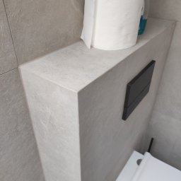 Firma ogolnobudowlana Jarzmik Kamil - Szara, wykafelkowana zabudowa stelaża WC z czarnym przyciskiem spłukującym, rolką papieru toaletowego i butelką płynu na górze. Widoczny fragment muszli toaletowej.