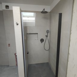 Firma ogolnobudowlana Jarzmik Kamil - Nowoczesna kabina prysznicowa z czarnymi profilami i deszczownicą, szare płytki na ścianach, widoczne niedokończone prace wykończeniowe.