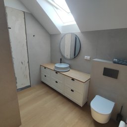 Firma ogolnobudowlana Jarzmik Kamil - Nowoczesna łazienka w Porąbce Iwkowska z umywalką nablatową, okrągłym lustrem i podwieszaną toaletą, częściowo wykończona, z widoczną surową ścianą przed wykończeniem.