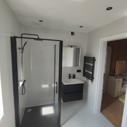 Firma ogolnobudowlana Jarzmik Kamil - Nowoczesna łazienka z czarną kabiną prysznicową typu walk-in, umywalką z szafką w tym samym kolorze oraz grzejnikiem drabinkowym, całość utrzymana w jasnej kolorystyce z akcentami imitacji marmuru...
