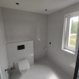 Firma ogolnobudowlana Jarzmik Kamil - Nowoczesna łazienka z białymi płytkami imitującymi marmur, wisząca toaleta z czarnym przyciskiem spłukiwania, czarne akcesoria i okno z widokiem na zieleń.