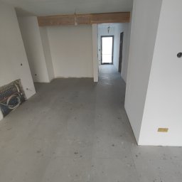 Firma ogolnobudowlana Jarzmik Kamil - Surowe wnętrze w trakcie prac wykończeniowych: szare płytki podłogowe, białe ściany z widocznymi instalacjami, drewniane elementy konstrukcyjne pod sufitem, widok na korytarz.
