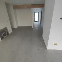 Surowe wnętrze w trakcie prac wykończeniowych: szare płytki podłogowe, białe ściany z widocznymi instalacjami, drewniane elementy konstrukcyjne pod sufitem, widok na korytarz.