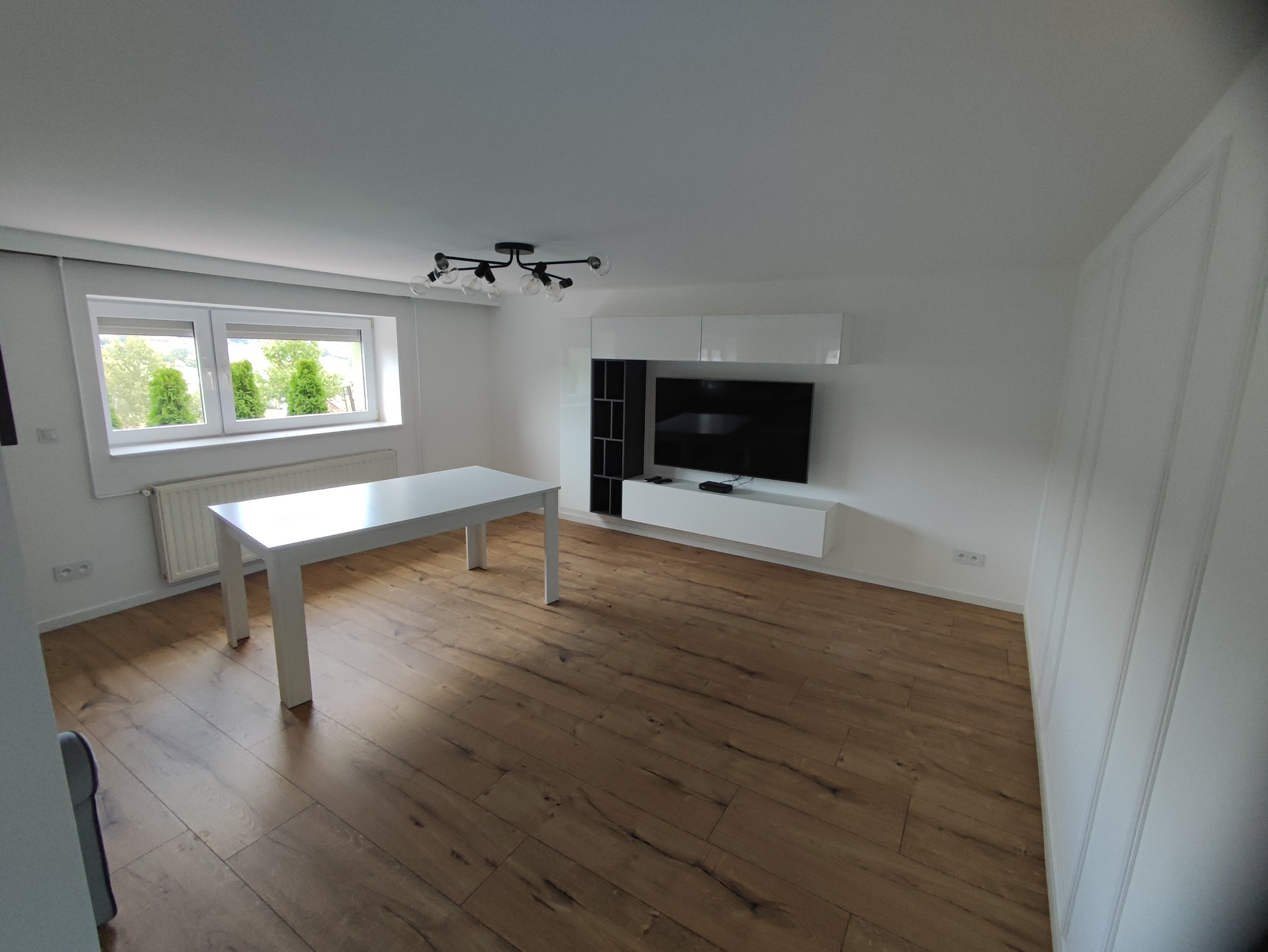 Jasne, przestronne wnętrze po remoncie z drewnianą podłogą, białym stołem, ścianą telewizyjną i nowoczesnym oświetleniem sufitowym.