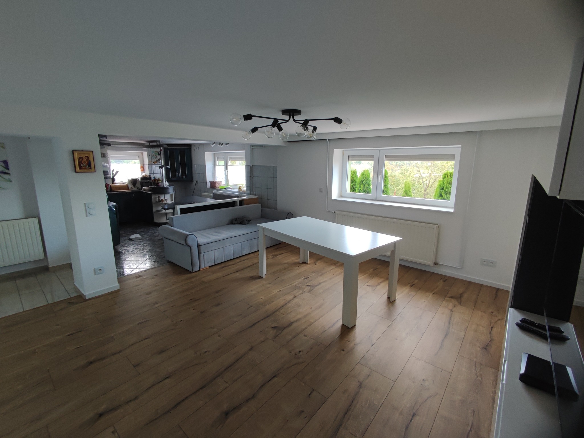 Salon z drewnianą podłogą, białym stołem i szarą sofą, widok w stronę kuchni z nowoczesną lampą sufitową i oknami z widokiem na zieleń.