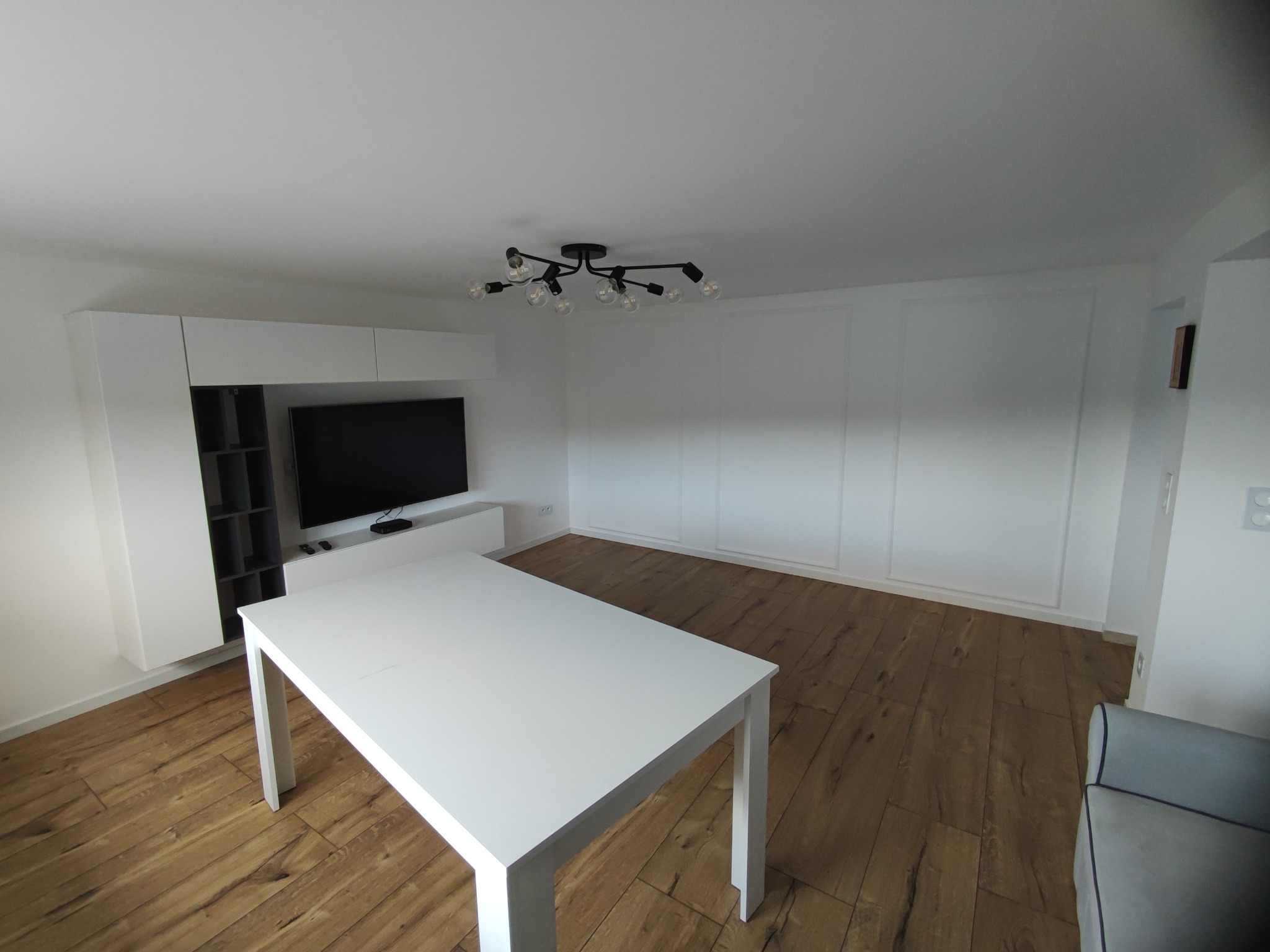 Jasne wnętrze salonu z drewnianą podłogą, białym stolikiem, zabudową TV i nowoczesną lampą sufitową z wieloma żarówkami.