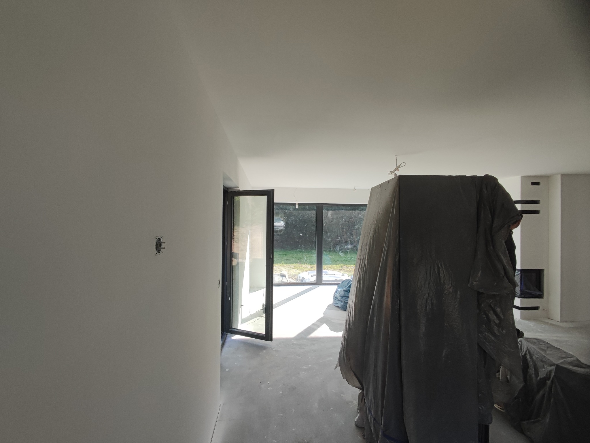 Wnętrze w trakcie prac przygotowawczych: ściany po gładzi, zabezpieczone meble folią, otwarte drzwi balkonowe.